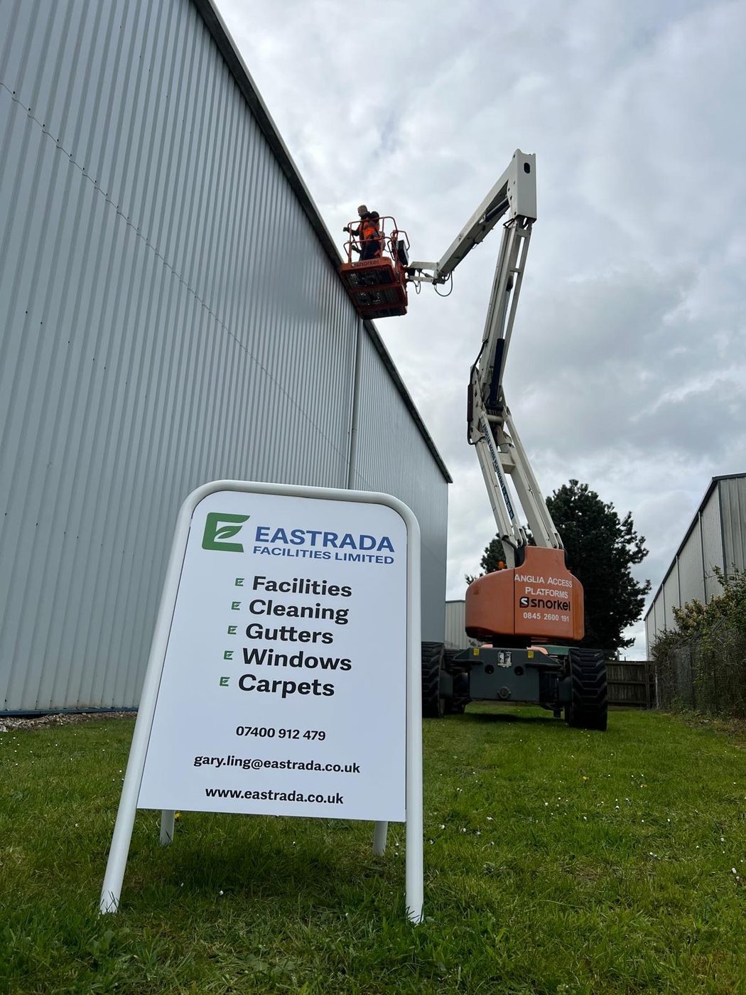 Eastrada Facilities team cleaning a warehouse exterior using a Snorkel cherry picker, with branded A-frame sign in foreground listing services: Facilities, Cleaning, Gutters, Windows, Carpets